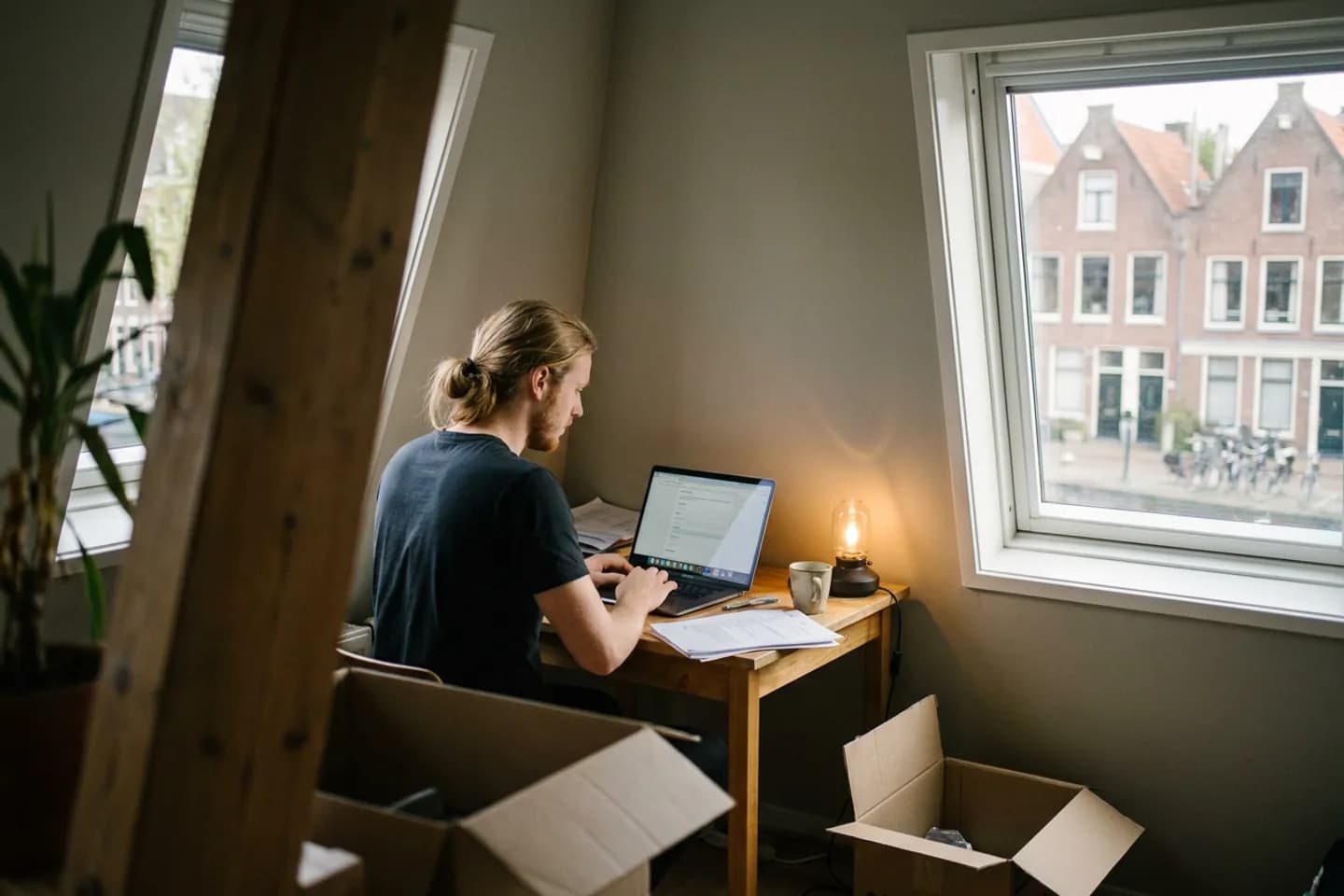 Een startende webontwikkelaar vult een online formulier in op zijn laptop in een net ingerichte zolderkamer, met dozen op de vloer.