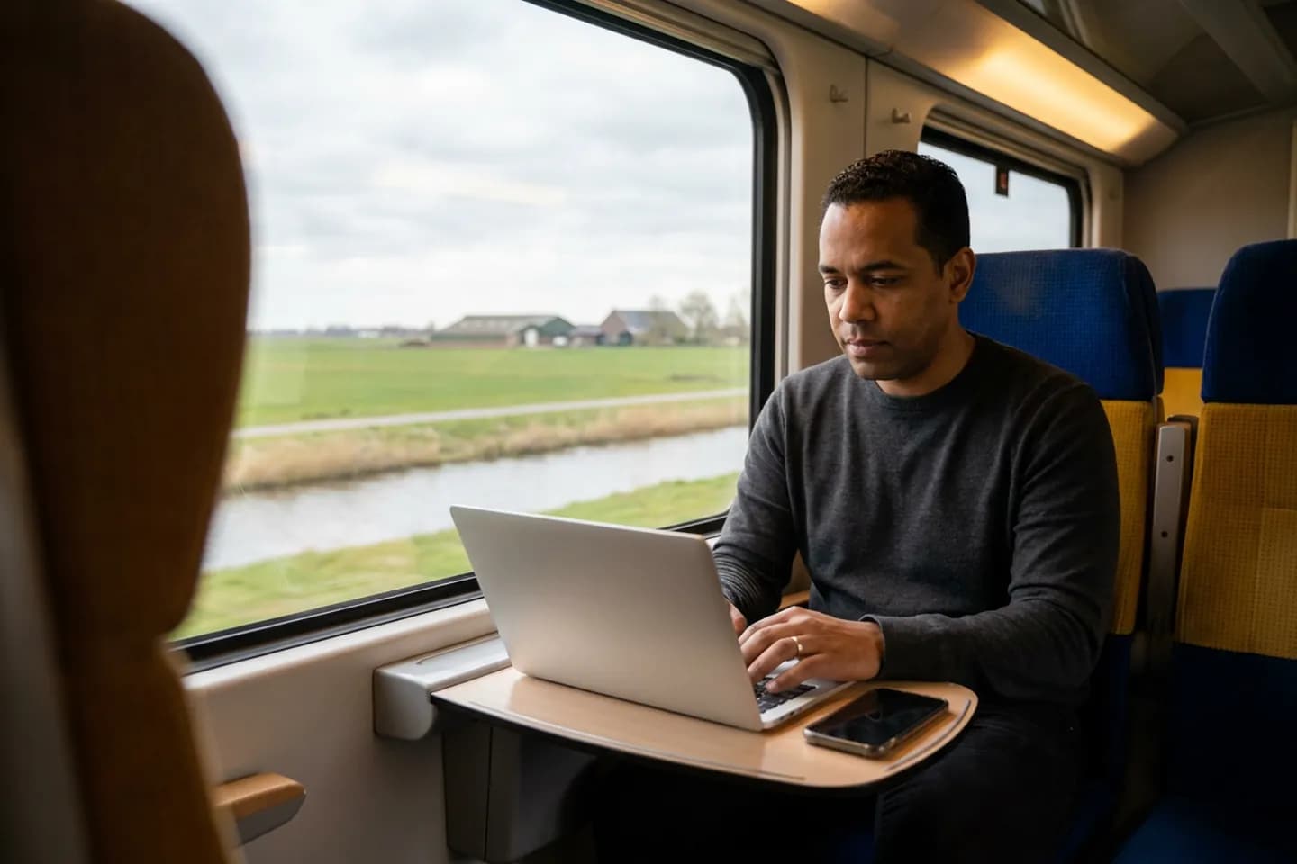 Een copywriter werkt op een laptop aan het klaptafeltje van een trein, terwijl buiten weilanden en een sloot voorbijkomen.