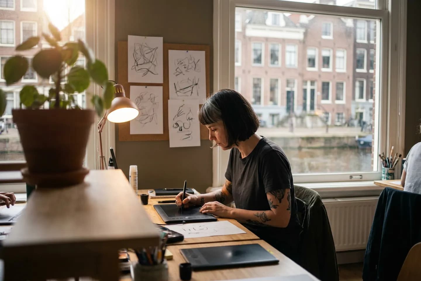 Een illustrator tekent op een tekentablet aan een bureau in een gedeelde studio, met schetsen op een prikbord.