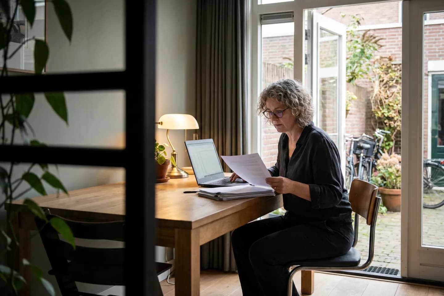 Een juridisch adviseur vergelijkt een papieren factuur met haar laptop aan een tafel naast de openstaande tuindeur.