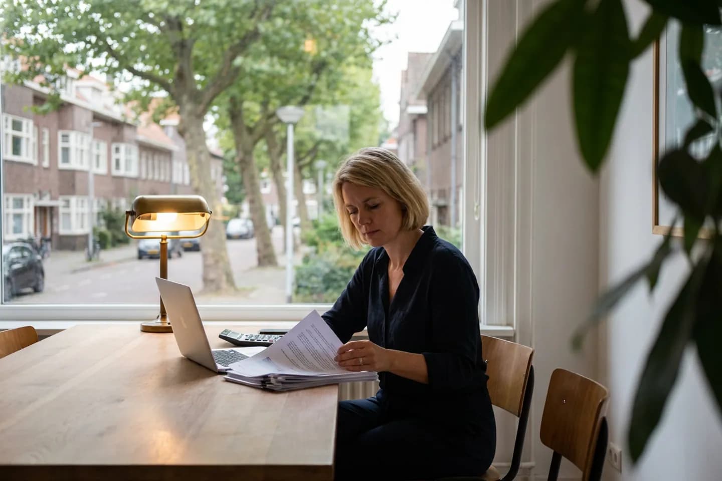 Een marketingconsultant bekijkt een geprinte offerte naast haar laptop en rekenmachine, aan een tafel voor een raam met uitzicht op een straat met bomen.
