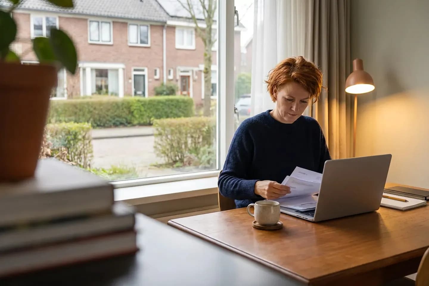 Een tekstschrijver bekijkt papieren facturen naast haar laptop en een kop koffie, aan een bureau voor een raam met uitzicht op een woonstraat.