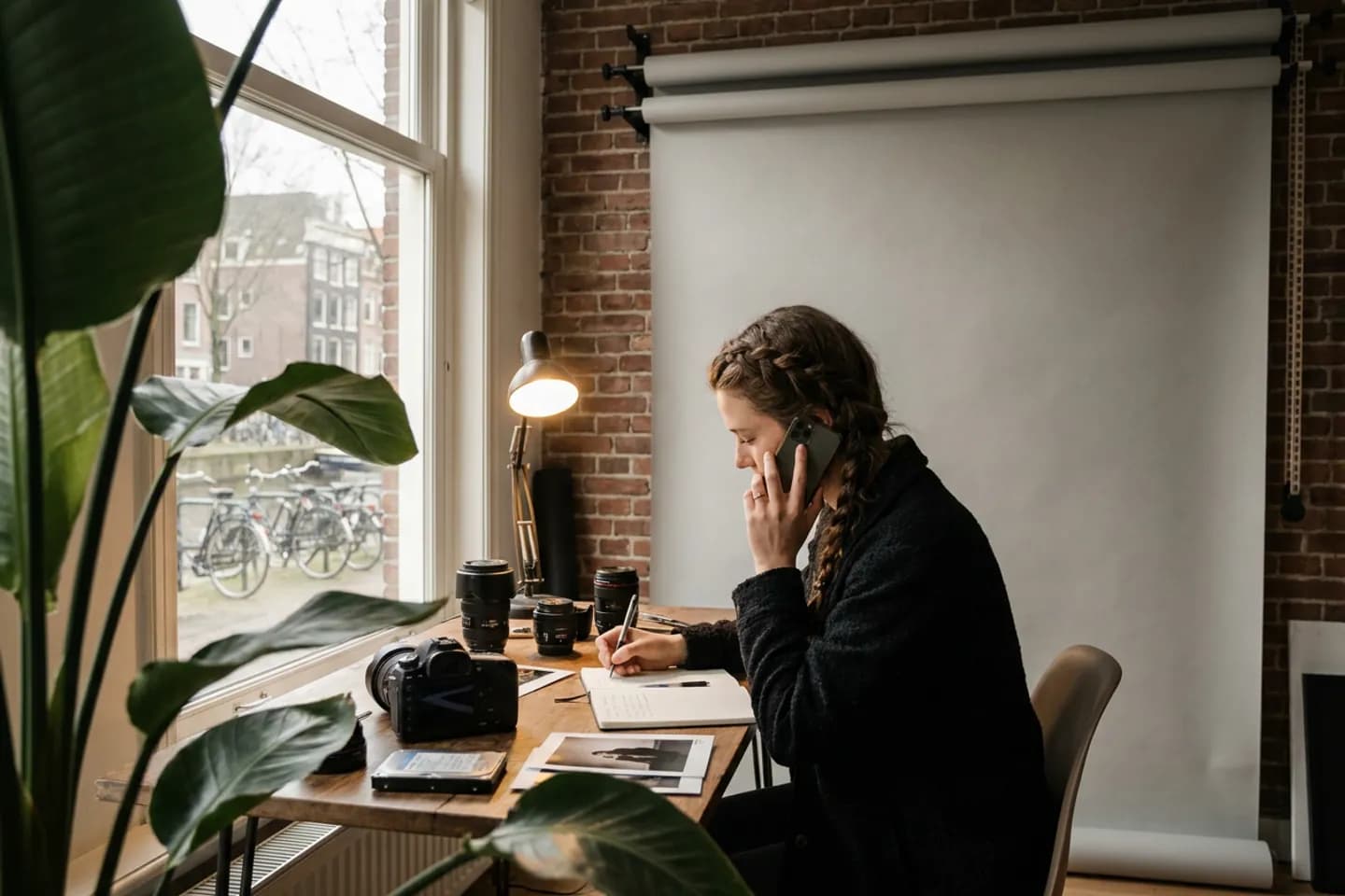 Een fotograaf belt en maakt een aantekening aan haar bureau in een thuisstudio, met een camera en lenzen voor zich.