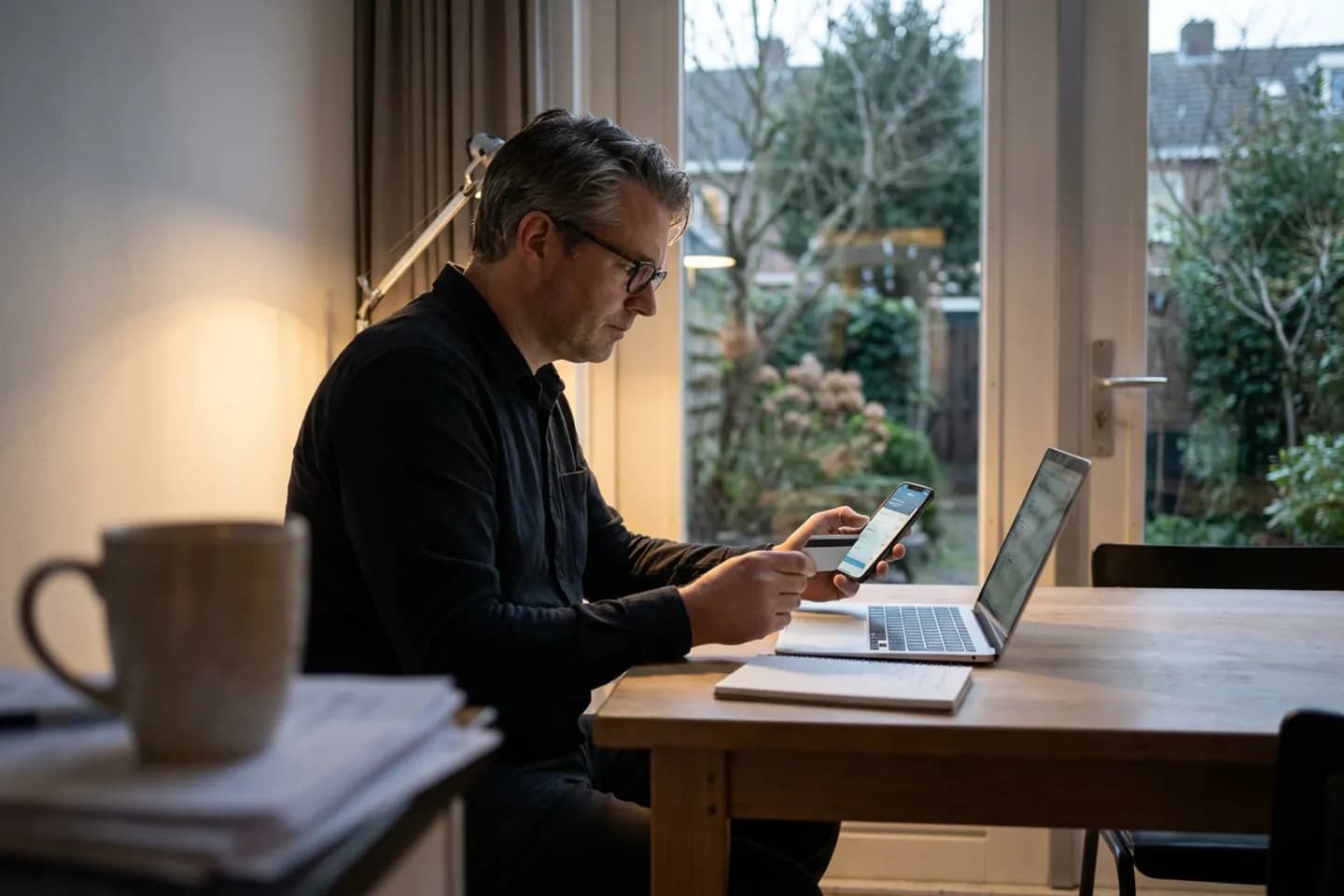 Een consultant kijkt op zijn telefoon aan de keukentafel, met een betaalpas en een laptop voor zich en de tuin achter hem.