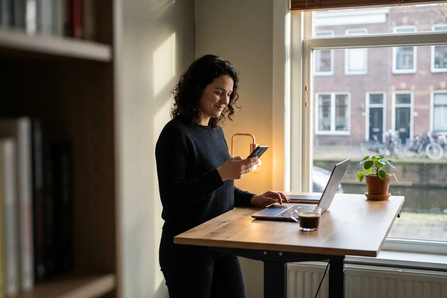 Een softwareontwikkelaar kijkt glimlachend op haar telefoon bij een sta-bureau voor het raam van haar thuiskantoor.