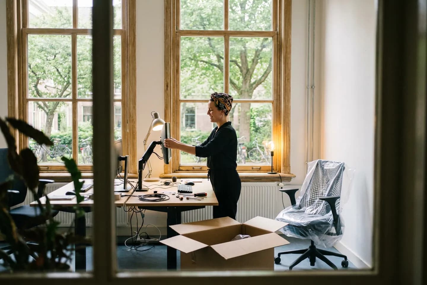 Een ontwerper zet een nieuw beeldscherm op haar bureau in een net gehuurde werkruimte, met een uitgepakte doos op de vloer en een stoel nog in plastic.
